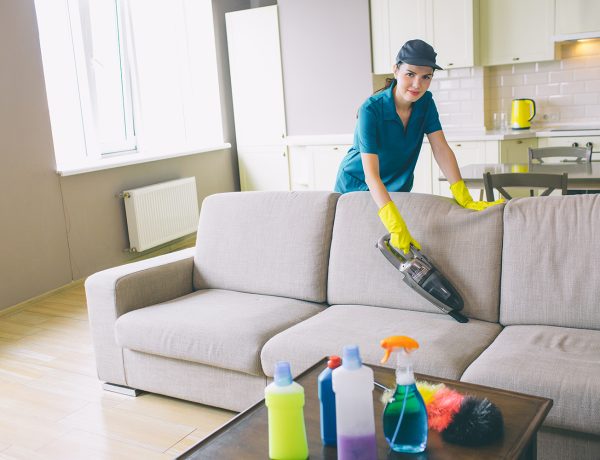Girl in unform stands behind sofa and cleans it with small vacuum cleaner. There are bottles with different liquid and rags on table